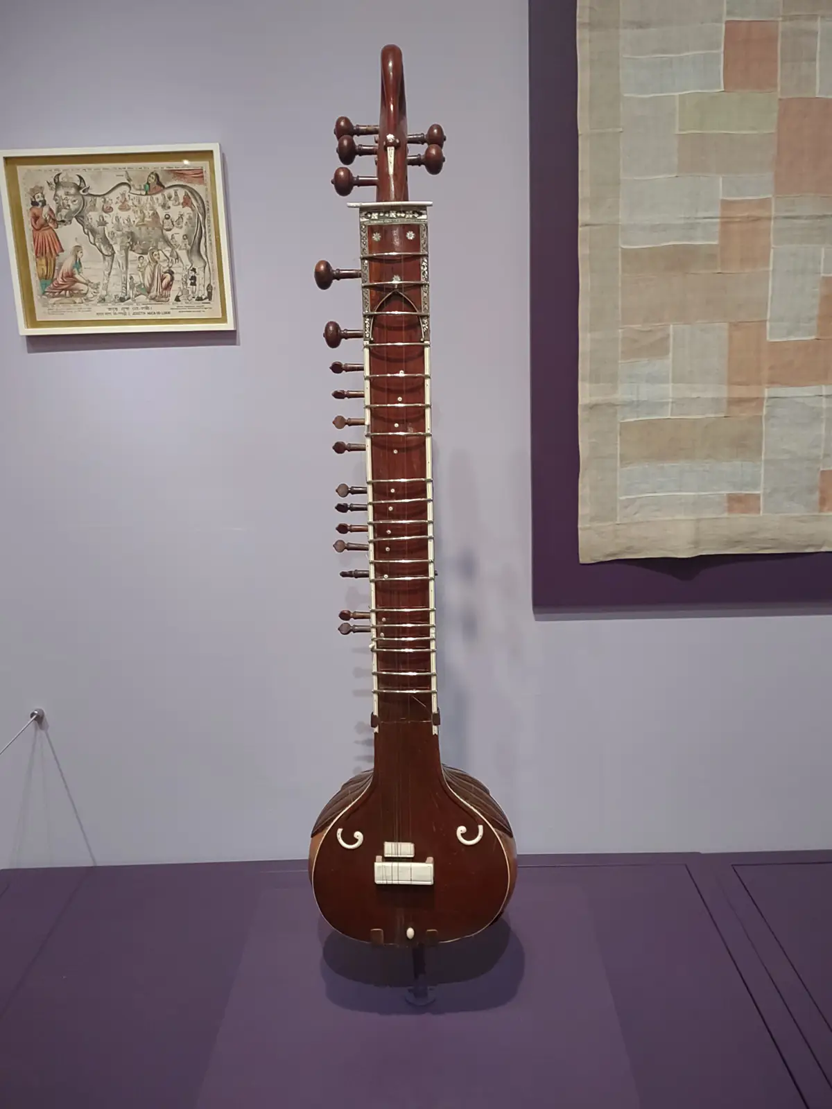 Museum gallery display of a tall sitar standing upright, flanked by a framed Indian folk painting and a patchwork textile.