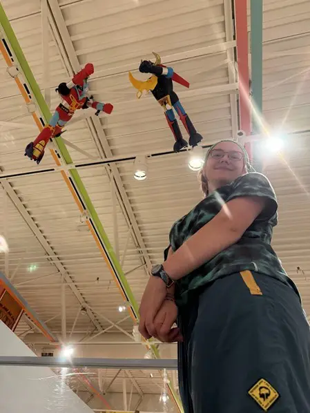 Flynn standing under two colorful vintage robot toys suspended from a warehouse-style ceiling with string lights.