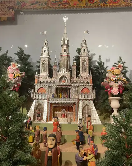 Silver Gothic szopka castle display with carved figures inside and flanking floral arrangements amid small evergreen trees.