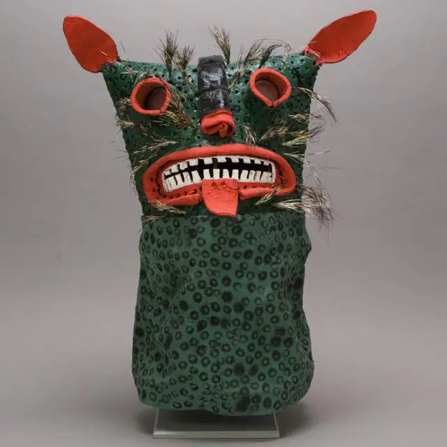 Green textured folk mask with pointed red ears, bared teeth, protruding tongue, bristled fur accents, and spotted fabric hood.