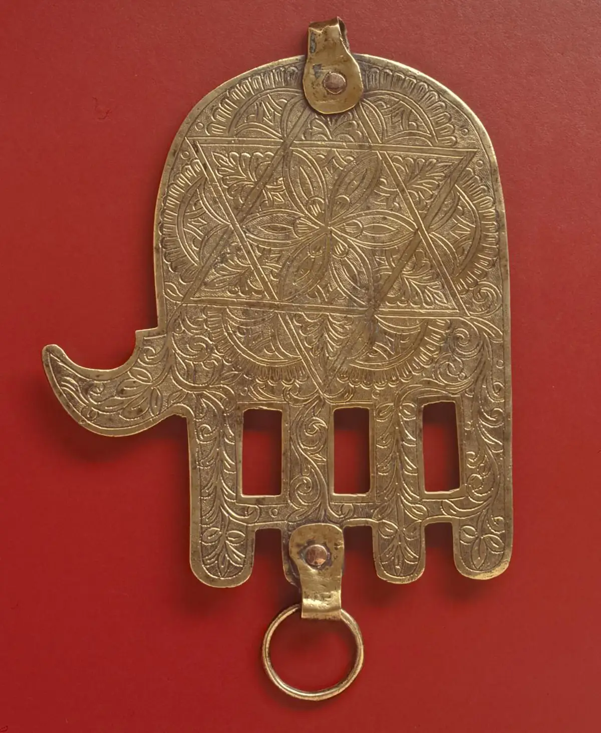 Gold-toned Hamsa hand amulet with geometric engravings and a small ring at the bottom, displayed on a red background.
