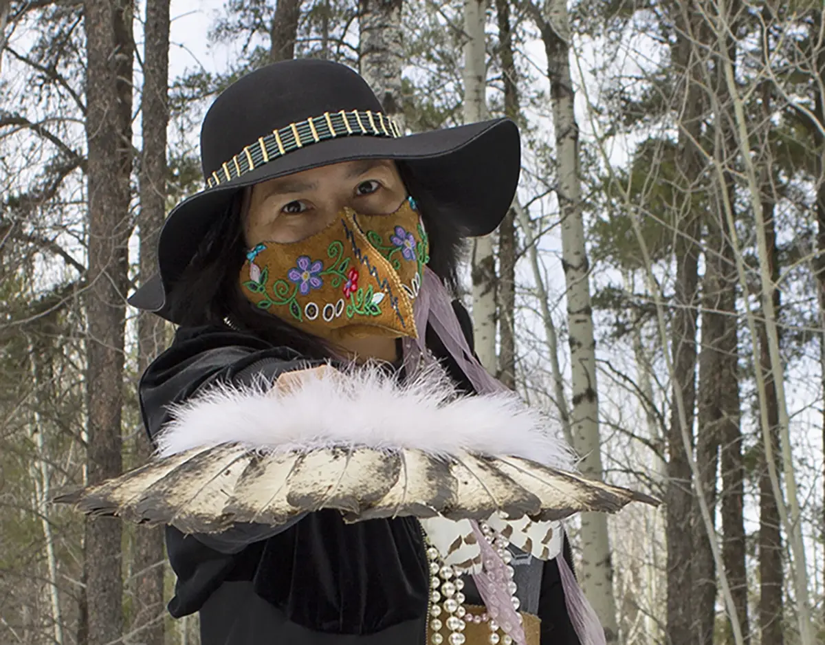 Woman wearing a wide-brimmed black hat, colorful floral face mask and fur-trimmed cloak standing among Aspen trees, holding a row of feathers out to the camera.
