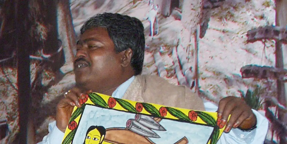 Man holding up a scroll that depicts a Bengali folktale about British colonialism, it shows a man's head on the front of a plane and falling, bullet shaped objects.